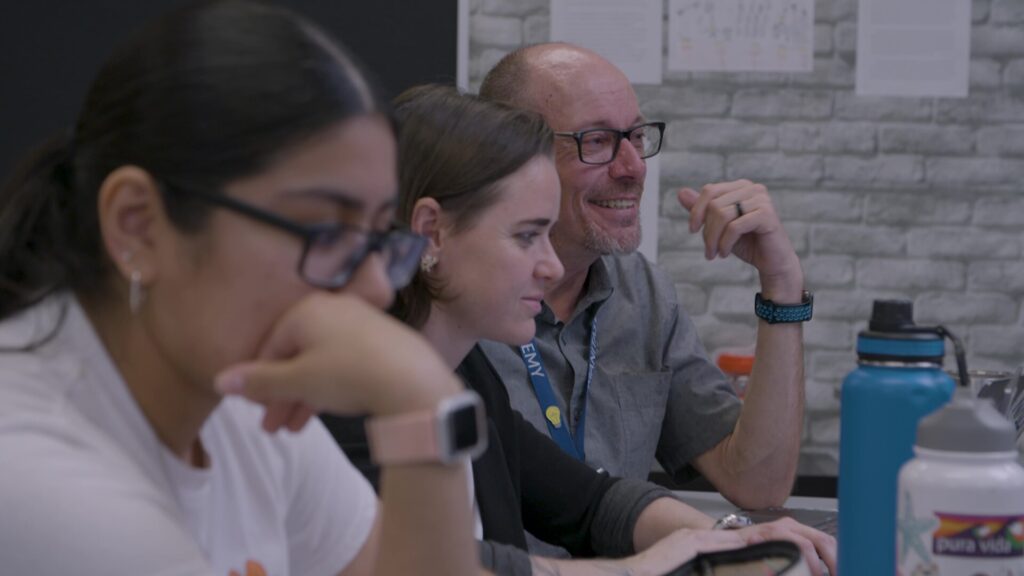 Two women and a man sit at a table, discussing and looking down at laptops.