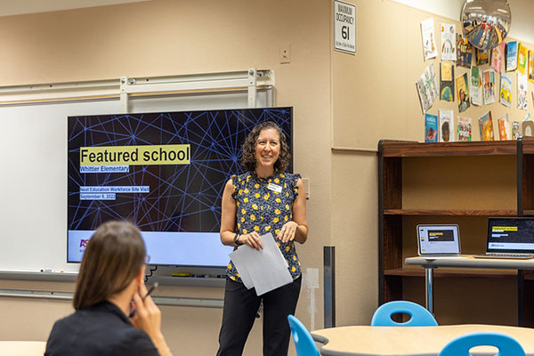 A woman stands in front of a screen featuring a PowerPoint with the text "Featured school."