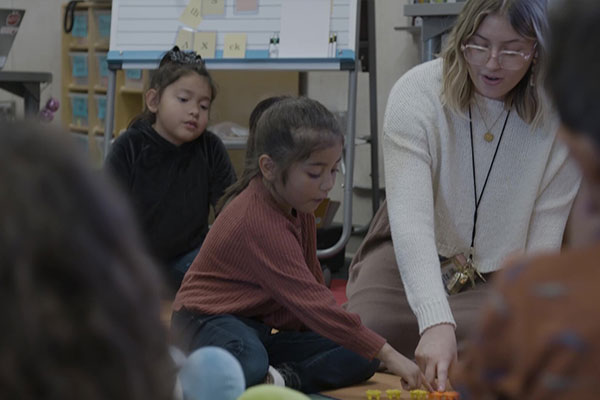 A woman in a white sweater sits with students on the floor.