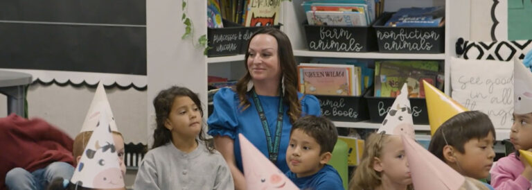 A woman wearing a blue top sits amongst a group of young kids in a classroom.