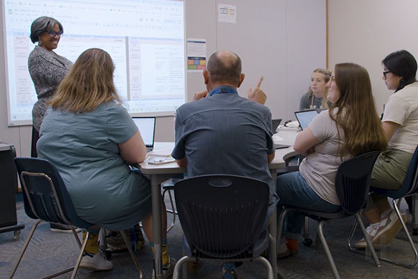 A woman stands in front of a group of educators in a classroom, smiling. 