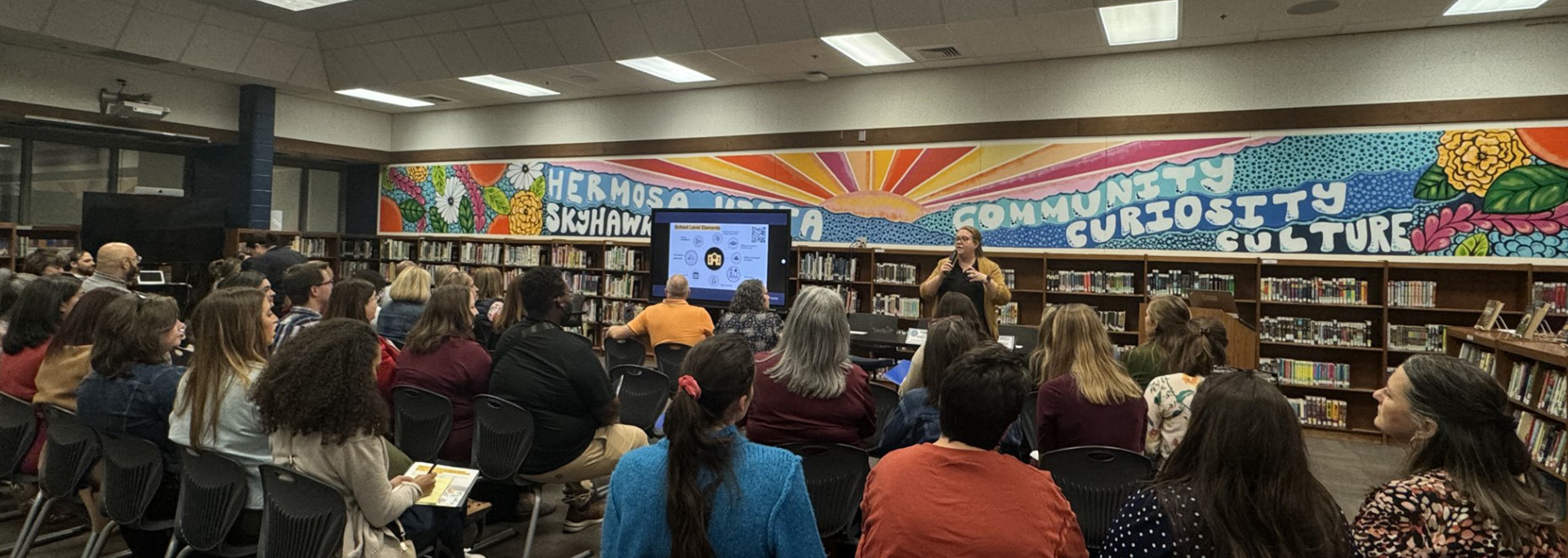 An image of a group of people sitting in a library watching a woman talk. Behind the woman is a colorful mural featuring a sunset and the words: Hermosa Vista Skyhawks on one side and Community, Curiosity, Culture on the other side