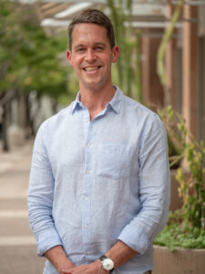 A man wearing a light blue button-down shirt smiles for a headshot.