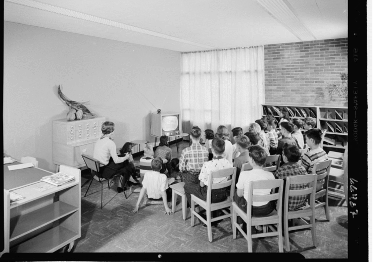 An old photo in black and white showing students sitting watching a TV in a library.